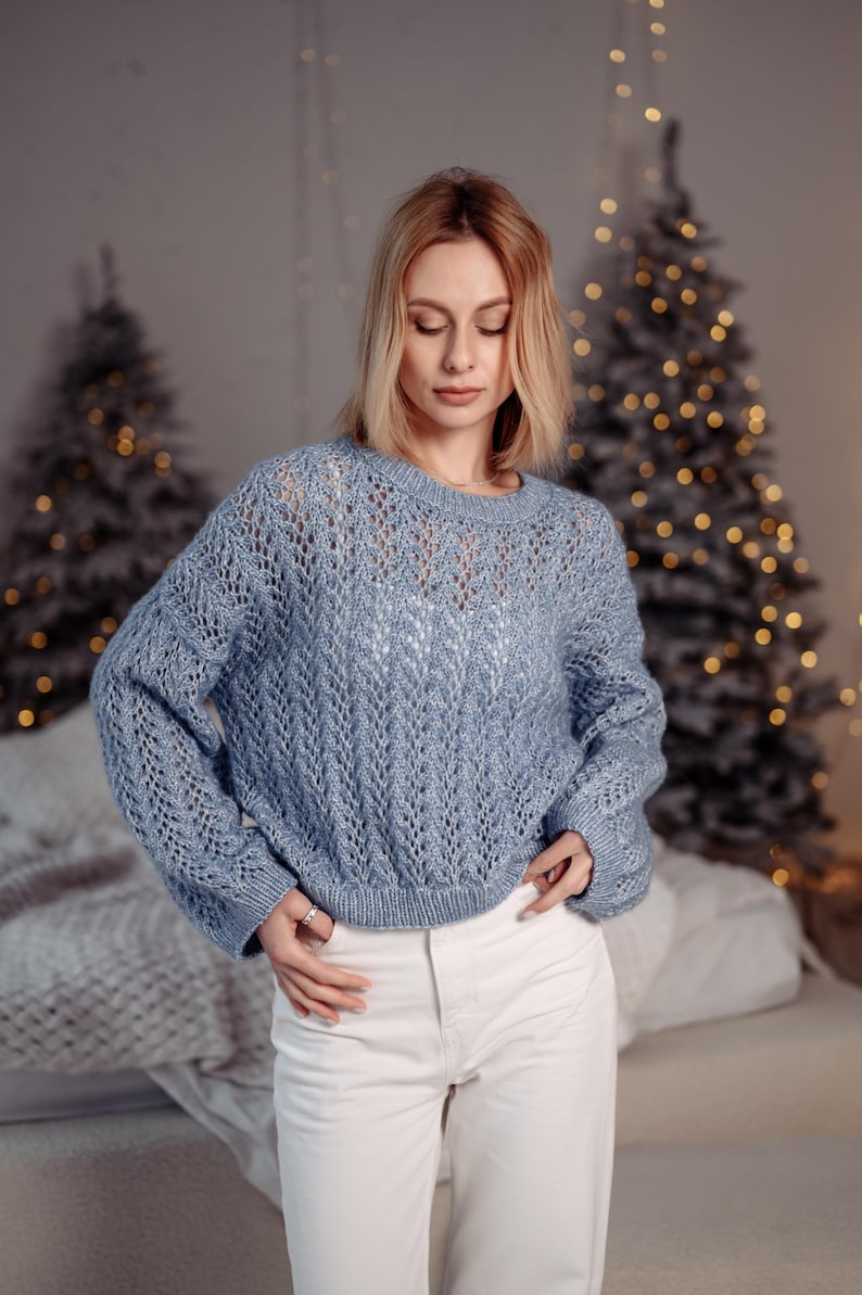 May include: A woman wearing a light blue knitted sweater with a delicate openwork pattern and white pants. She is standing in front of a Christmas tree with lights.