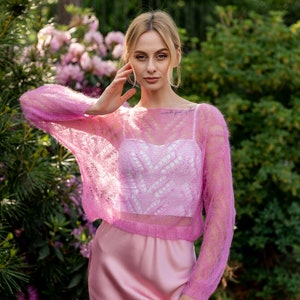 May include: A woman wearing a pink, lacy, knitted sweater over a white tank top and a pink satin skirt. She is standing in a garden with pink flowers.