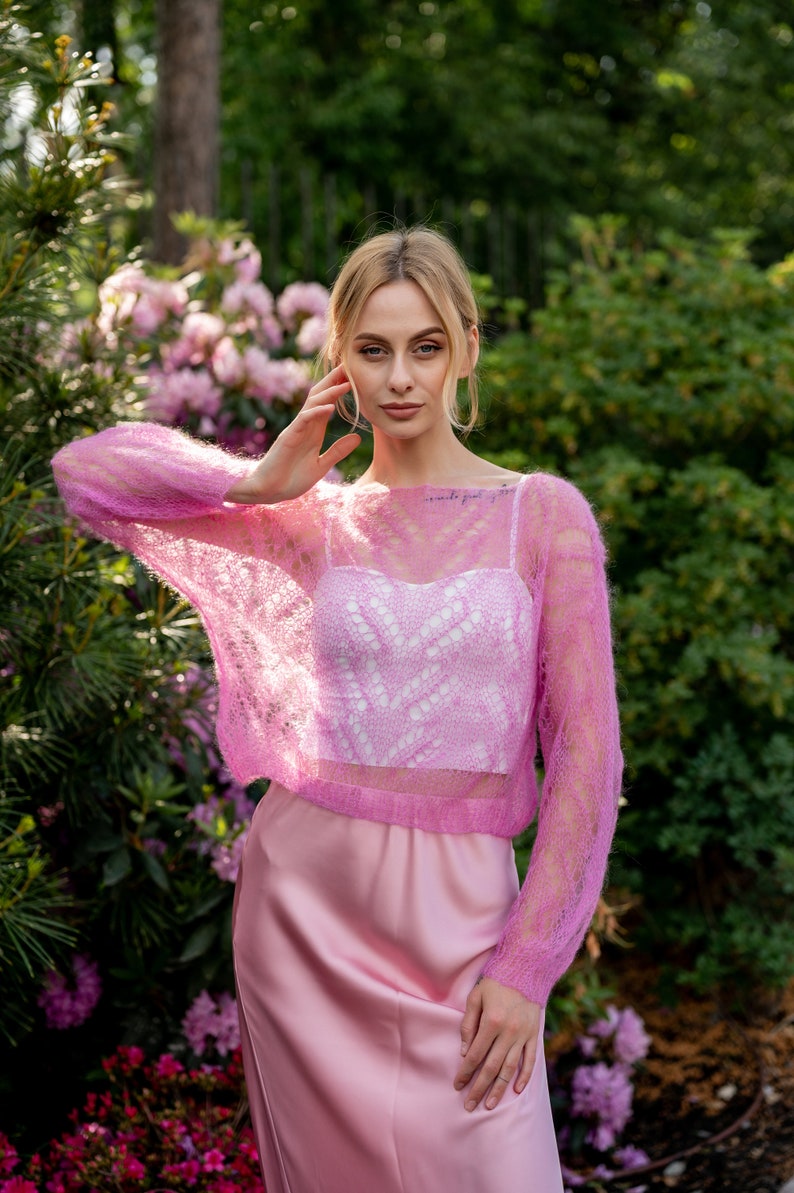 May include: A woman wearing a pink, lacy, knitted sweater over a white tank top and a pink satin skirt. She is standing in a garden with pink flowers.