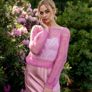 May include: A woman wearing a pink, sheer, knitted sweater with a pink satin skirt. She is standing in front of a bush of pink flowers.