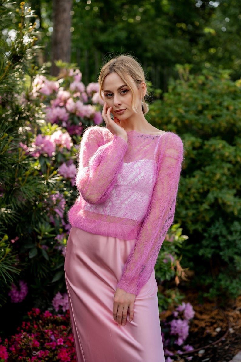 May include: A woman wearing a pink, sheer, knitted sweater with a pink satin skirt. She is standing in front of a bush of pink flowers.