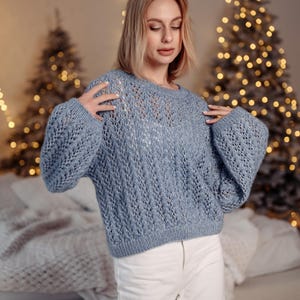 May include: A woman wearing a light blue knitted sweater with a delicate openwork pattern. The sweater has long sleeves and a round neckline. She is standing in front of a Christmas tree with lights.