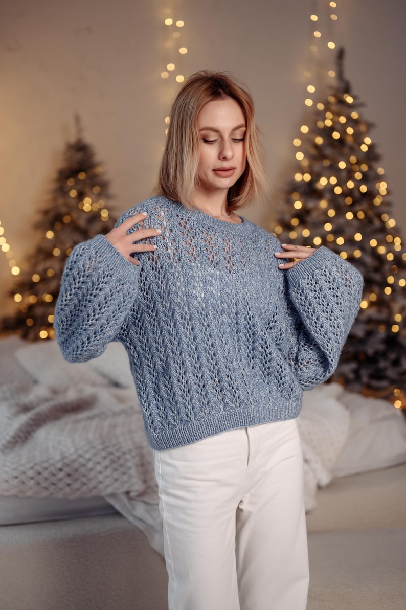 May include: A woman wearing a light blue knitted sweater with a delicate openwork pattern. The sweater has long sleeves and a round neckline. She is standing in front of a Christmas tree with lights.