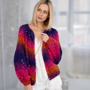 May include: A colourful, open-front cardigan in a gradient of colours, transitioning from deep purple to bright pink, with orange and red accents. The knitwear is embellished with sequins. The sleeves are long and voluminous.