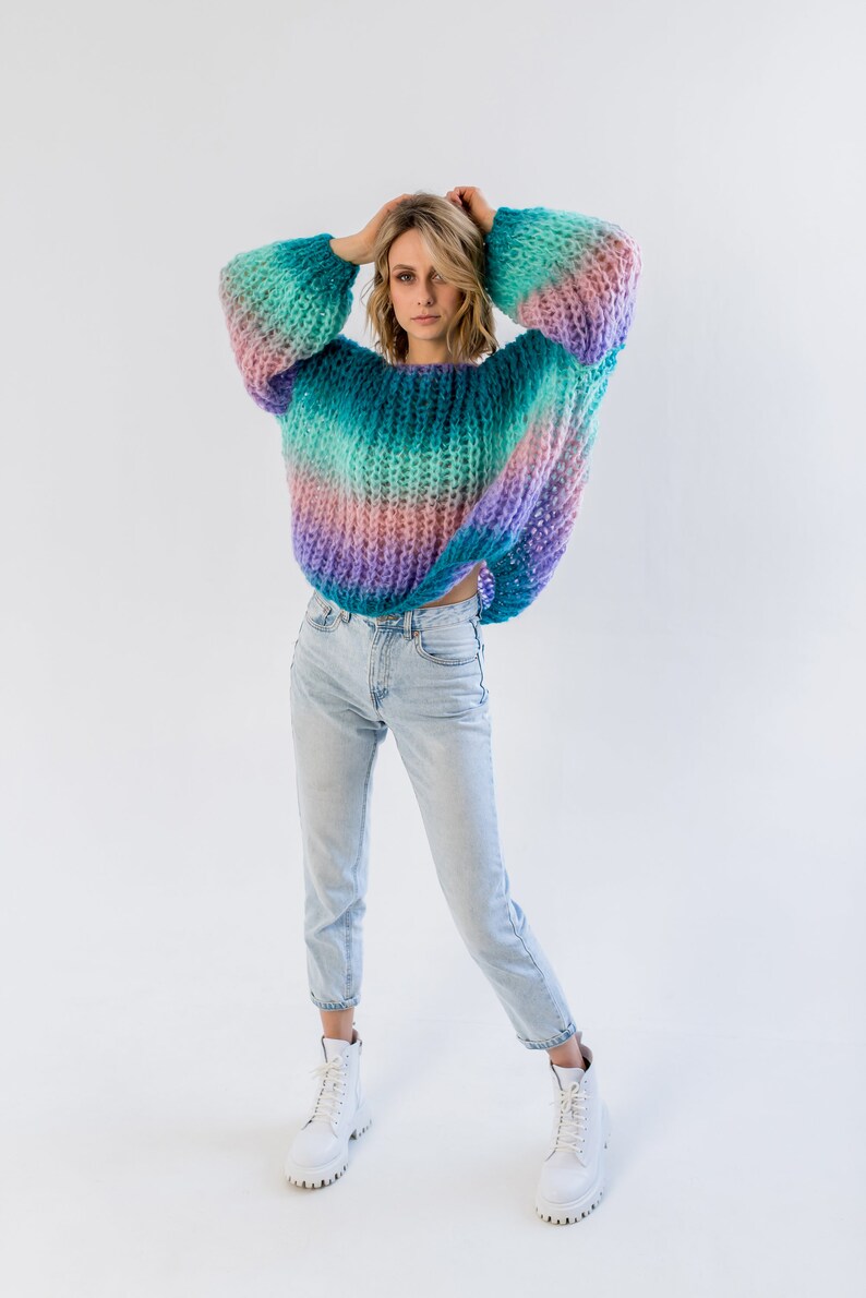 Może przedstawiać: Kobieta w kolorowym, paskowanym swetrze z dzianiny, jasnoniebieskich dżinsach i białych butach sportowych.