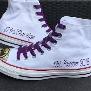 Puede incluir: Zapatillas blancas de lona de ca&ntilde;a alta con cordones morados. La zapatilla izquierda tiene el texto "Mrs Claridge" bordado en el lateral. La zapatilla derecha tiene un logotipo de Batman negro y amarillo con pedrer&iacute;a y el texto "12th October 2018" bordado en el lateral.