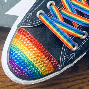 May include: Black canvas Converse sneakers with a rainbow-colored rhinestone design on the toe and rainbow striped laces.