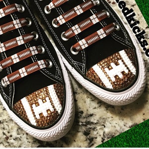 May include: Black Converse sneakers with brown and white football-themed shoelaces and brown and white rhinestones on the toe. The shoes have the text "CONVERSE" on the side. The text "www.trickedkicks.com" is visible in the background.