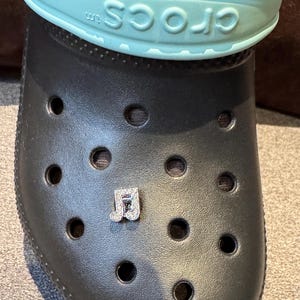 May include: A black clog with a blue strap that says "crocs tm" on it. A small charm with the letters "JD" in rhinestones is attached to the clog.