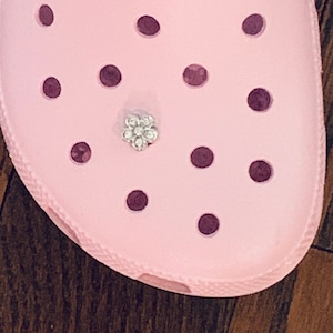 May include: A pink shoe with a flower-shaped rhinestone charm. The shoe has a textured surface and multiple holes.