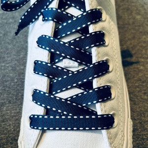 May include: Close-up of white sneakers with navy blue shoelaces with white stitching. The shoelaces are tied in a criss-cross pattern.