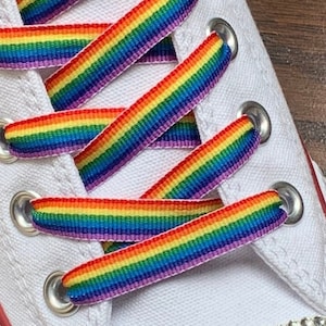 May include: Rainbow shoelaces tied on a pair of white canvas shoes. The shoelaces are made of a woven fabric with alternating stripes of red, orange, yellow, green, blue, indigo, and violet.