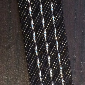 May include: A close-up of a black beaded bracelet with silver accents. The bracelet is made of small, round beads that are strung together in a tight pattern. The silver accents are small, shiny beads that are interspersed throughout the black beads.