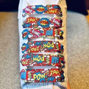 Puede incluir: Un par de zapatillas blancas con cordones coloridos que presentan palabras de estilo cómic como "POW", "ZAP" y "BANG".