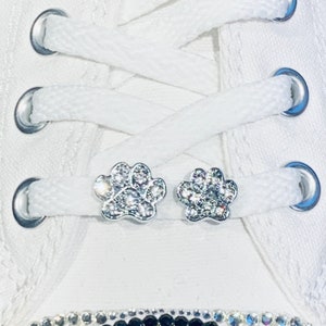 May include: Two silver paw print shoe charms with clear rhinestones. The charms are attached to white shoelaces.
