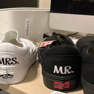 May include: A pair of white and black canvas slip-on shoes with the words "MRS." and "MR." printed on them. The shoes are a popular style for weddings.