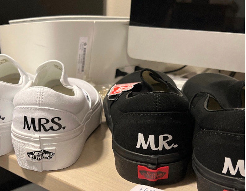 May include: A pair of white and black canvas slip-on shoes with the words "MRS." and "MR." printed on them. The shoes are a popular style for weddings.