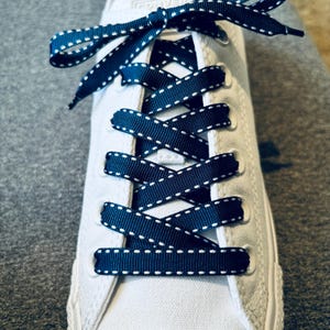 May include: White sneakers with navy blue shoelaces that have white stitching. The shoelaces are tied in a criss-cross pattern.