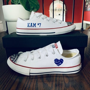 May include: White Converse sneakers with blue and red accents. The left shoe has the text "KAM #7" on the side. The right shoe has a blue volleyball heart design.