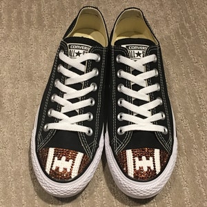 May include: Black Converse sneakers with white laces and a brown and white football design on the toe. The football design is made of rhinestones.