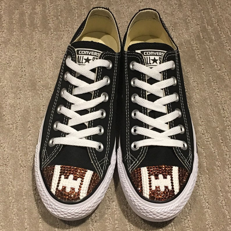 May include: Black Converse sneakers with white laces and a brown and white football design on the toe. The football design is made of rhinestones.