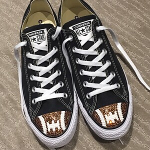 May include: Black Converse sneakers with white laces and a brown and white football design on the toe. The football design is covered in rhinestones.