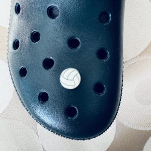 May include: A navy blue clog shoe with a white and gray volleyball charm. The shoe has multiple round holes and a textured surface. The volleyball charm is round with black lines.