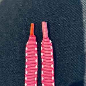 Puede incluir: Dos cordones de zapatos rosas con lunares blancos. Un cordón tiene una punta roja y el otro una punta transparente.