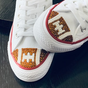 May include: White canvas shoes with a red trim and brown rhinestones forming a football design on the toe.