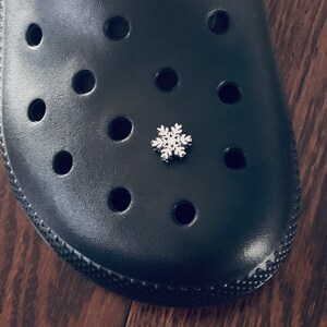 May include: A black rubber shoe with a snowflake charm attached to one of the holes. The shoe has a textured surface and multiple holes.