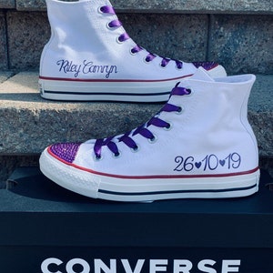 Puede incluir: Zapatillas Converse blancas de ca&ntilde;a alta con cordones de cinta morada. La zapatilla izquierda tiene los nombres "Riley Camryn" bordados en el lateral. La zapatilla derecha tiene la fecha "26&hearts;10&hearts;19" bordada en el lateral. Las zapatillas est&aacute;n sobre una caja de zapatos Converse negra.