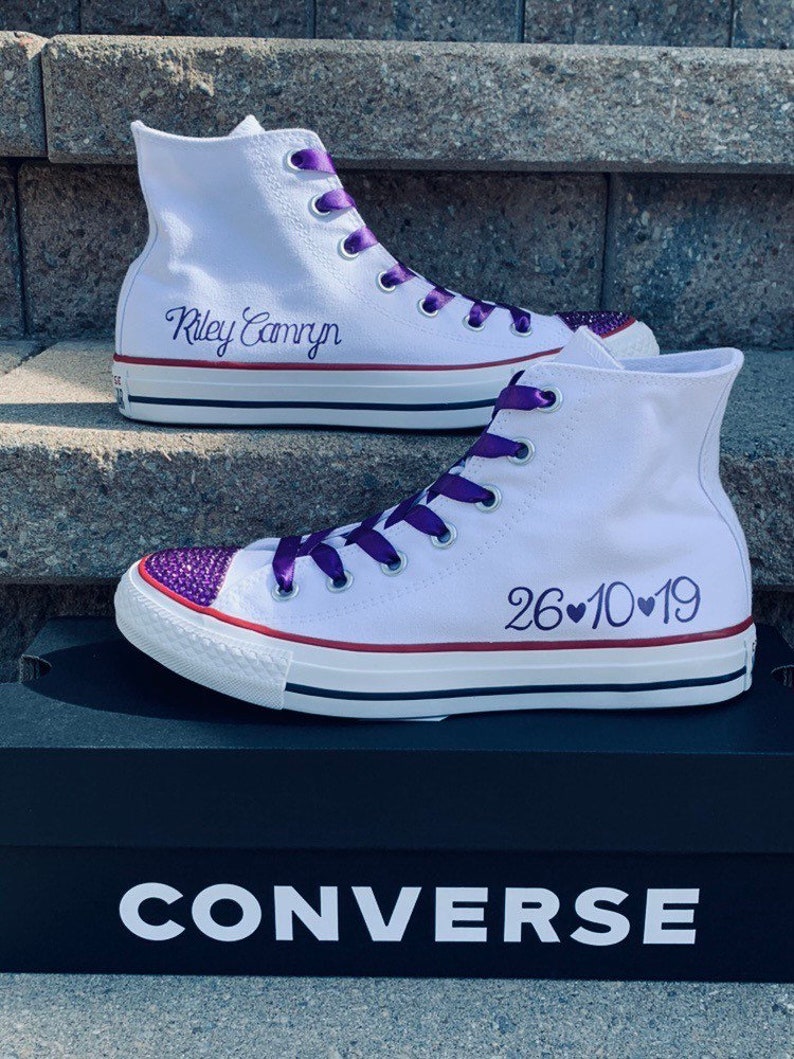 Puede incluir: Zapatillas Converse blancas de ca&ntilde;a alta con cordones de cinta morada. La zapatilla izquierda tiene los nombres "Riley Camryn" bordados en el lateral. La zapatilla derecha tiene la fecha "26&hearts;10&hearts;19" bordada en el lateral. Las zapatillas est&aacute;n sobre una caja de zapatos Converse negra.