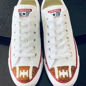 May include: White Converse All Star sneakers with a red trim and a brown and white football design on the toe. The football design is made of rhinestones.