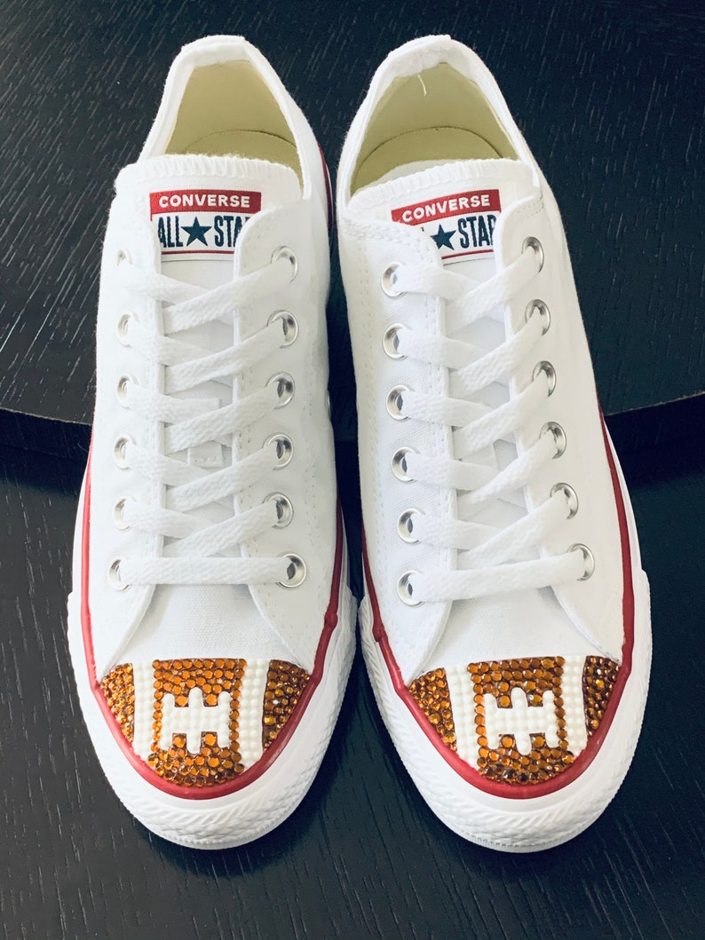 May include: White Converse All Star sneakers with a red trim and a brown and white football design on the toe. The football design is made of rhinestones.