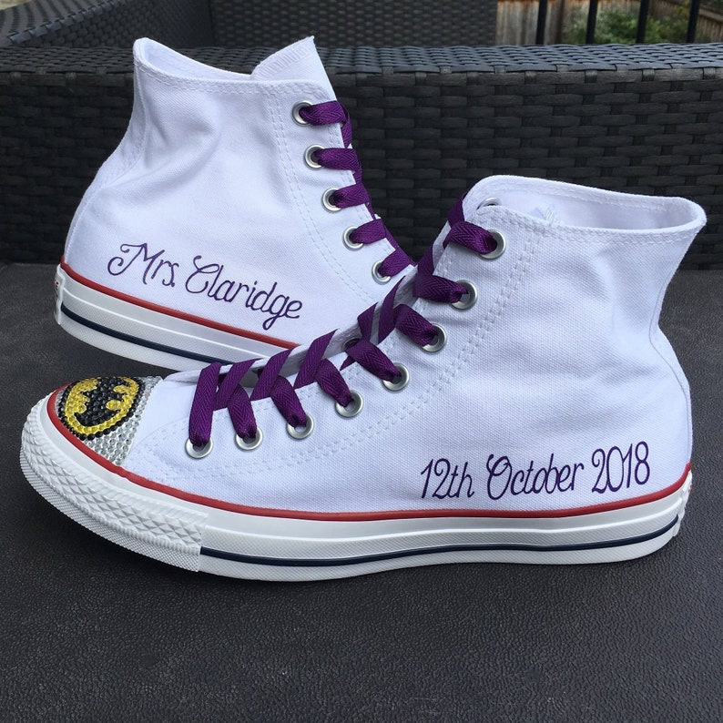 Puede incluir: Zapatillas blancas de lona de ca&ntilde;a alta con cordones morados. La zapatilla izquierda tiene el texto "Mrs Claridge" bordado en el lateral. La zapatilla derecha tiene un logotipo de Batman negro y amarillo con pedrer&iacute;a y el texto "12th October 2018" bordado en el lateral.