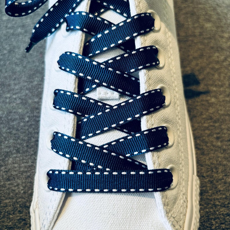 May include: Close-up of white sneakers with navy blue shoelaces with white stitching. The shoelaces are tied in a criss-cross pattern.