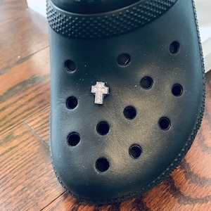May include: A black clog with a small, silver cross charm with clear rhinestones attached to the top of the shoe.