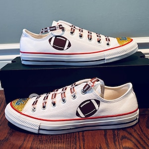 May include: White canvas low-top sneakers with brown and white striped laces. The shoes have a brown football design on the side and gold glitter on the toe.