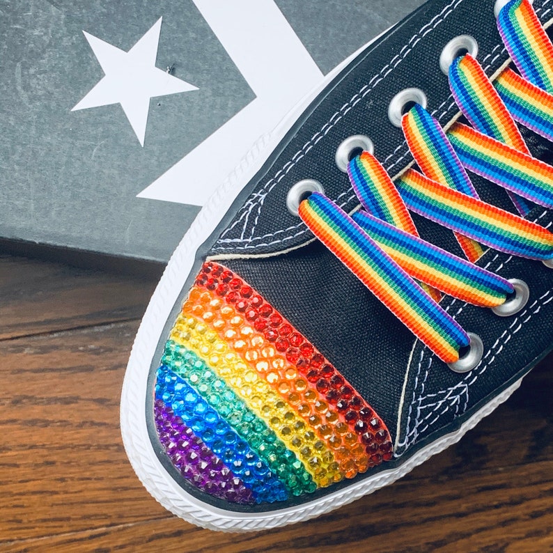 May include: Black canvas Converse sneakers with a rainbow-colored rhinestone design on the toe and rainbow striped laces.