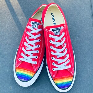 May include: Red Converse sneakers with a rainbow rhinestone design on the toe. The sneakers have white laces and the Converse logo is visible on the side.