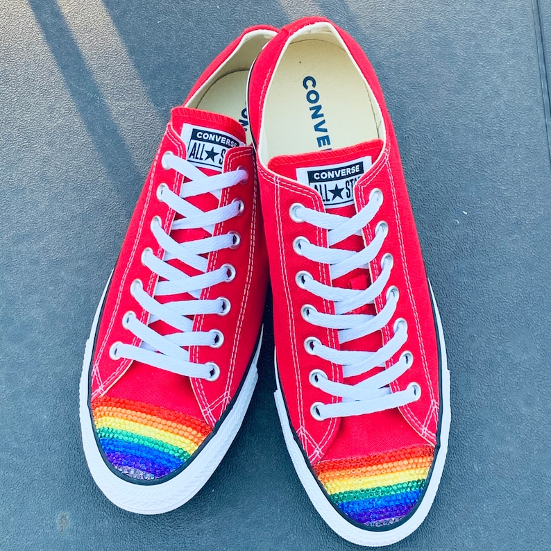 May include: Red Converse sneakers with a rainbow rhinestone design on the toe. The sneakers have white laces and the Converse logo is visible on the side.