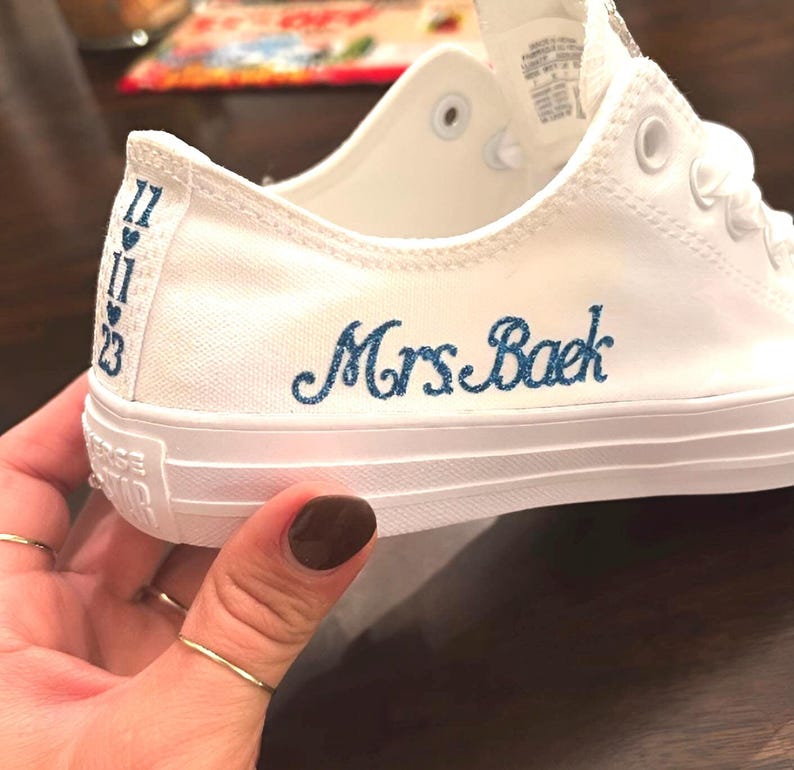 Puede incluir: Zapatillas blancas de lona con bordado azul que dice "Mrs. Baek" en el lateral. Las zapatillas tienen una suela de goma blanca y cordones blancos.