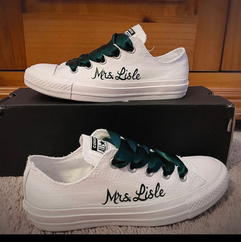 Puede incluir: Zapatillas blancas de lona con cordones de cinta de sat&eacute;n verde. Los zapatos est&aacute;n personalizados con el texto "Mrs. Lisle" en escritura negra.