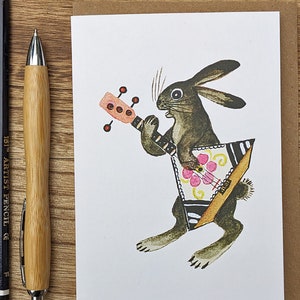 May include: A brown rabbit with a black and white striped tail plays a triangular musical instrument with a pink flower design. The instrument has a brown handle and black strings.