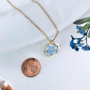 May include: A gold-colored chain necklace with a hexagonal pendant. The pendant contains a small, light blue flower preserved in clear resin. A small white pearl hangs next to the pendant. A one-cent coin is next to the necklace.