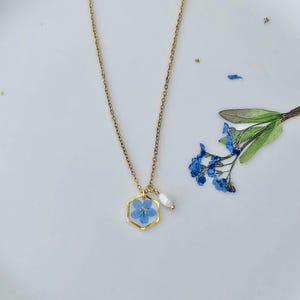 May include: A gold-colored necklace featuring a hexagonal pendant with a blue flower encased in clear resin. A small white pearl hangs next to the pendant. The necklace is displayed on a white surface with scattered blue flower petals.