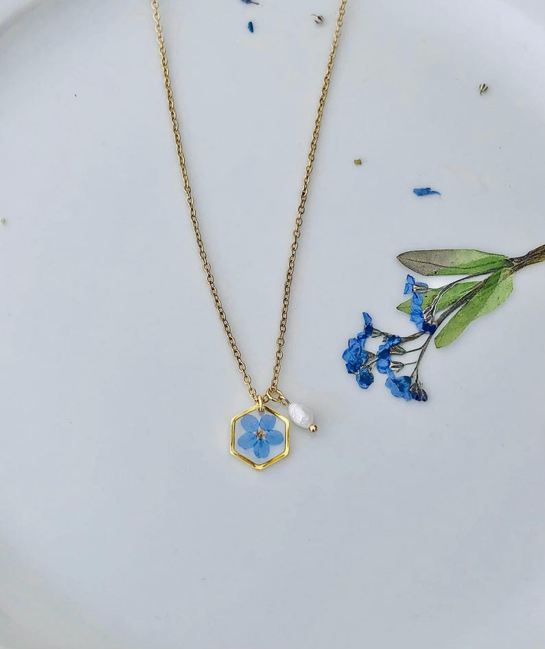 May include: A gold-colored necklace featuring a hexagonal pendant with a blue flower encased in clear resin. A small white pearl hangs next to the pendant. The necklace is displayed on a white surface with scattered blue flower petals.