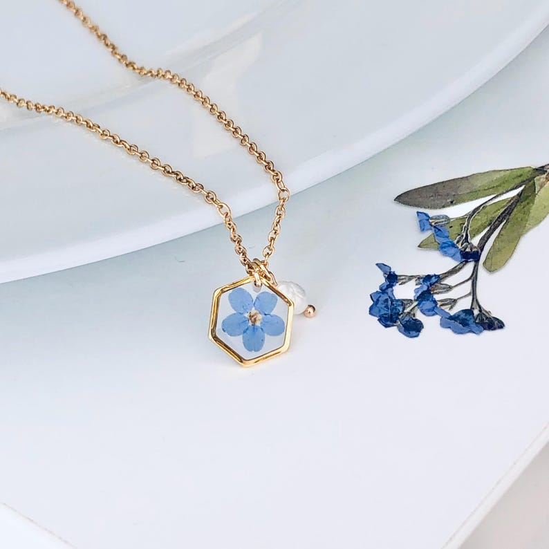 May include: A gold-colored necklace featuring a hexagonal pendant with a pressed blue flower encased in clear resin. A small white pearl charm hangs below the pendant. Additional blue flowers and green leaves are visible nearby.