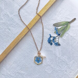 May include: A delicate gold-colored chain necklace with a hexagonal pendant. The pendant contains a small blue flower preserved in clear resin, with a small white pearl accent. The necklace is displayed on a white surface with a wooden accent and dried flowers.