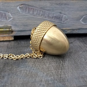 Pode incluir: Um colar com um pingente de bolota de ouro, com uma tampa texturizada detalhada e um corpo liso e arredondado. O colar é feito de uma delicada corrente de ouro.